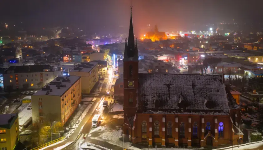 Nocna panorama Kościerzyny zimową porą. Widać ośnieżony kościół, oświetloną ulicę i kolorowe budynki. To piękne kościerzyna atrakcje.