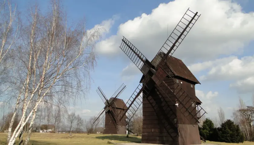 Na polanie stoi cmentarzysko wiatraków. Dwa drewniane wiatraki z wielkimi skrzydłami, na tle błękitnego nieba z chmurami.