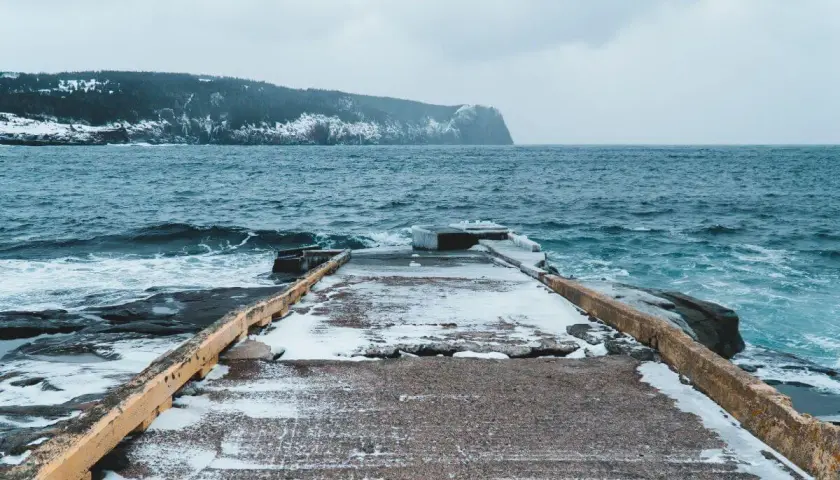 Zimowa sceneria nad morzem. Betonowe molo pokryte śniegiem, wzburzone, ciemne fale rozbijają się o skaliste brzegi. W oddali widać zalesione, ośnieżone wzgórza.