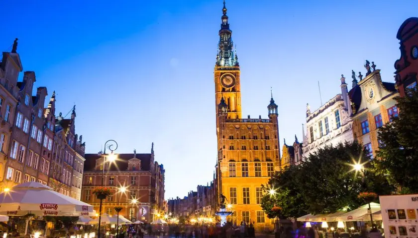 Wieczorny widok na Długi Targ, gdzie góruje ratusz Głównego Miasta w Gdańsku. Tętniące życiem kawiarnie i spacerujący ludzie.