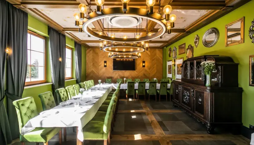 Elegancka sala bankietowa z długim stołem, zielonymi aksamitnymi krzesłami i stylowym, okrągłym żyrandolem. W tle zabytkowa komoda, a na ścianach ozdobne lustra. Idealne miejsce na spotkanie w stylu nordowi mol.
