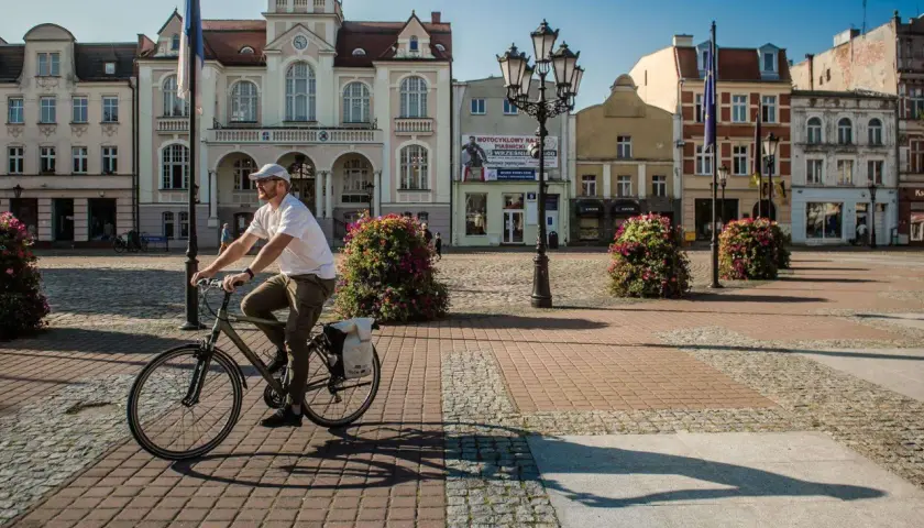 Mężczyzna na rowerze przejeżdża przez rynek przed ratuszem w Wejherowie. Słoneczny dzień, brukowana nawierzchnia, ozdobne krzewy.