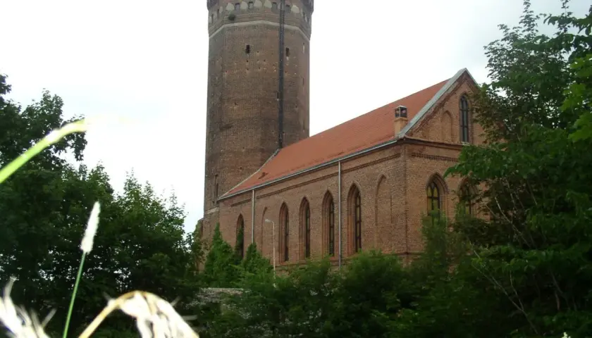 Zamek Człuchów, ceglana wieża i budynek z czerwonym dachem, otoczone zielenią drzew.