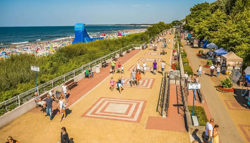 Tętniąca życiem ustka promenada w słoneczny dzień. Ludzie spacerują, odpoczywają na ławkach, a w tle widać plażę pełną turystów i morze.