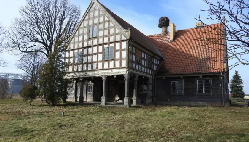 Stary dwór z muru pruskiego, z charakterystycznym gniazdem bociana na kominie. To historyczny lwi dwór, otoczony zielenią.