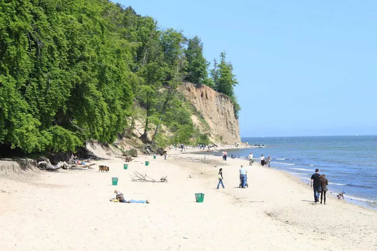 Piękny dzień na kąpieliskach Gdynia. Ludzie spacerują po piaszczystej plaży u podnóża zielonego klifu, ciesząc się słońcem i morzem.