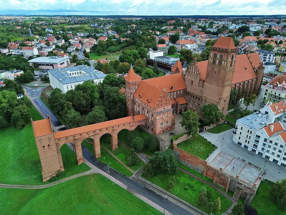 Zamek w Kwidzynie z charakterystycznym gdaniskiem i długim mostem nad fosą, otoczony zielenią i zabudowaniami miasta.