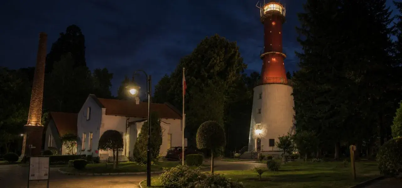 Wieczorem na przylądku na pobrzeżu kaszubskim widać oświetloną latarnię morską, budynki i ceglany komin.