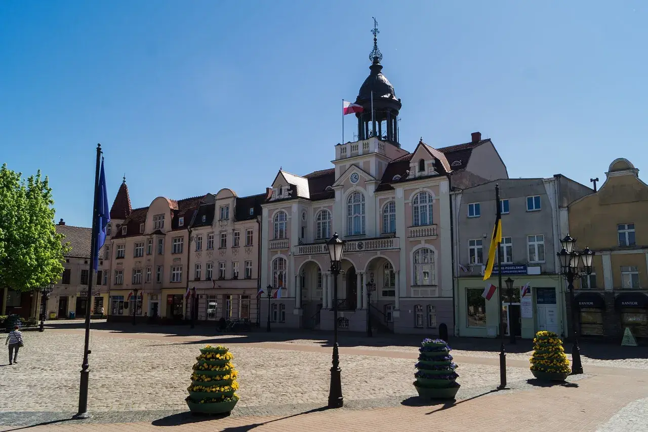 Słoneczny wejherowo rynek z ratuszem i kolorowymi budynkami. Na bruku stoją ozdobne donice z kwiatami.