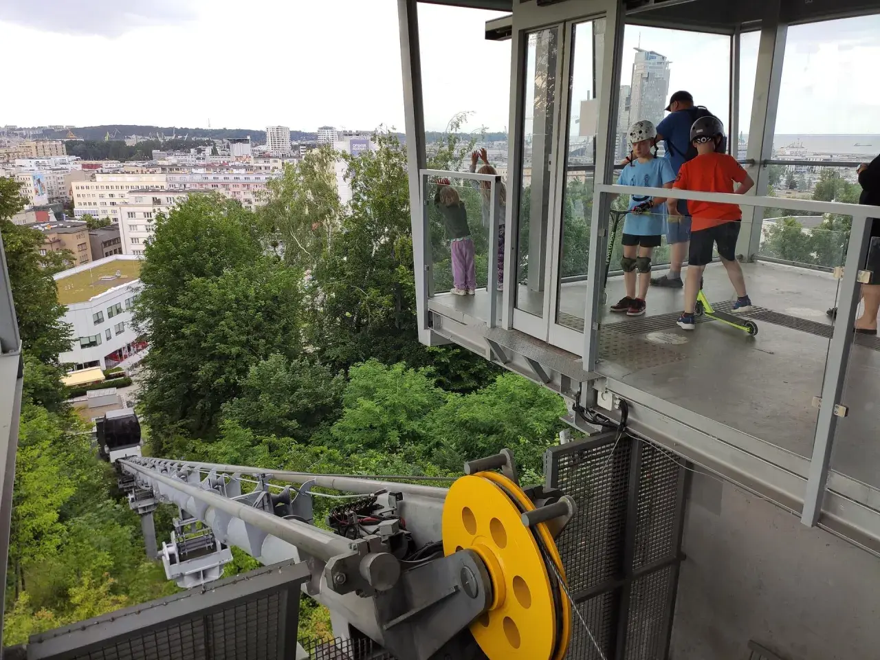 Dzieci na hulajnogach czekają na wjazd kolejką na kamienną górę w Gdyni, podziwiając panoramę miasta.