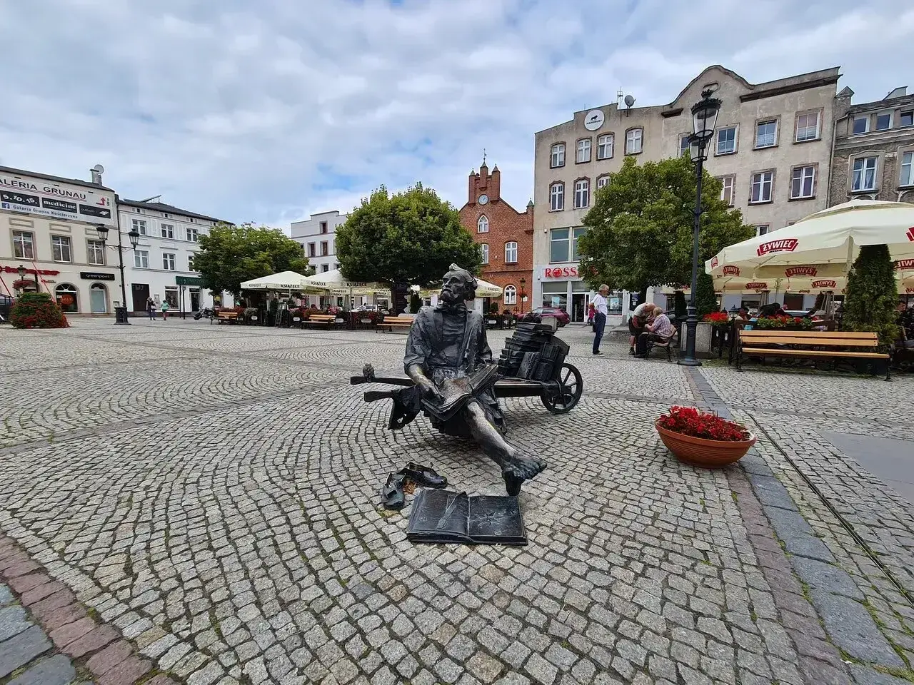 Pomnik pisarza na brukowanym kościerzyna rynek. W tle budynki, drzewa i ogródki kawiarniane.