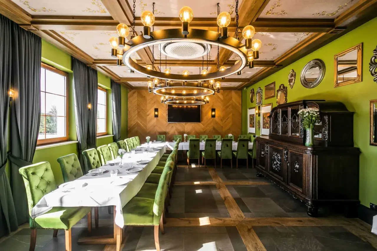 Elegancka sala bankietowa z długim stołem, zielonymi aksamitnymi krzesłami i stylowym, okrągłym żyrandolem. W tle zabytkowa komoda, a na ścianach ozdobne lustra. Idealne miejsce na spotkanie w stylu nordowi mol.