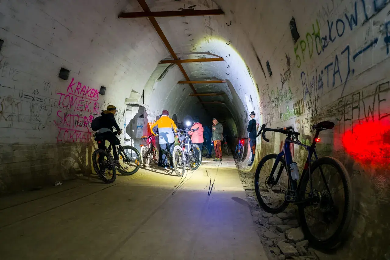 Grupa rowerzystów eksploruje mroczną, graffiti-pokrytą podziemną trasę rowerową, oświetloną latarkami.