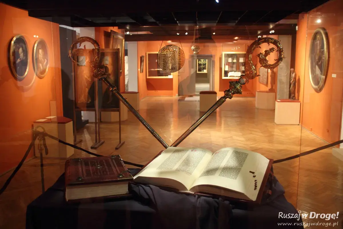 Otwarta biblia gutenberga pelplin, obok zamknięta księga i dwa ozdobne pastorały w muzeum.