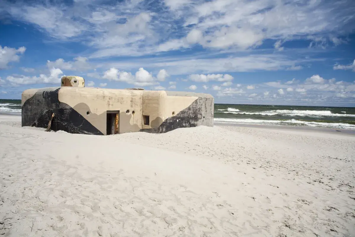 Betonowy bunkier z kamuflażem na piaszczystej plaży w Jastarni. Morze i niebo z chmurami w tle.