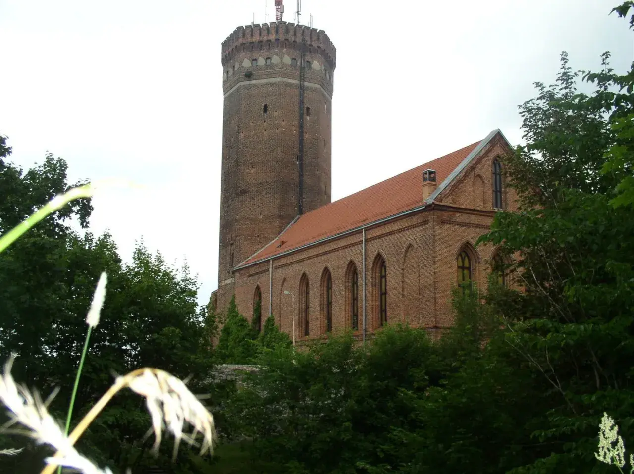 Zamek Człuchów, ceglana wieża i budynek z czerwonym dachem, otoczone zielenią drzew.