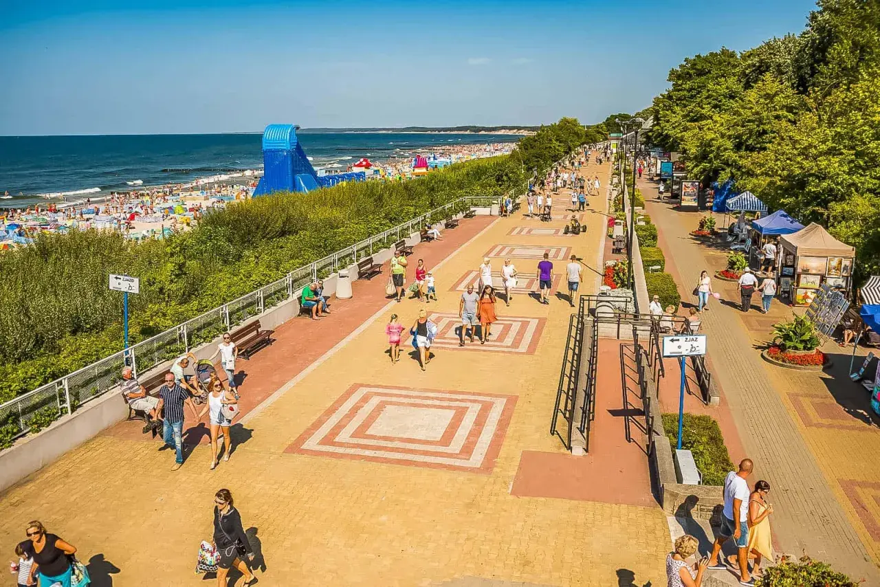 Tętniąca życiem ustka promenada w słoneczny dzień. Ludzie spacerują, odpoczywają na ławkach, a w tle widać plażę pełną turystów i morze.