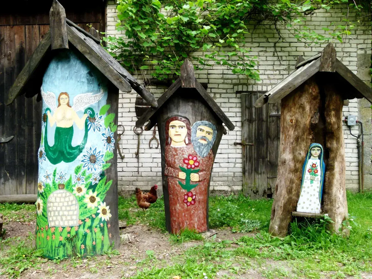 Trzy ozdobne ule, jeden z syreną, drugi z parą ludzi, trzeci z figurką Matki Boskiej. W tle kura i zielona roślinność. Brusy jaglie.