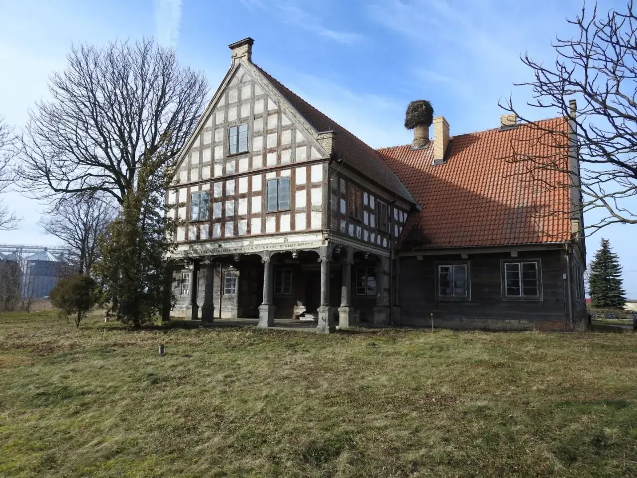 Stary dwór z muru pruskiego, z charakterystycznym gniazdem bociana na kominie. To historyczny lwi dwór, otoczony zielenią.