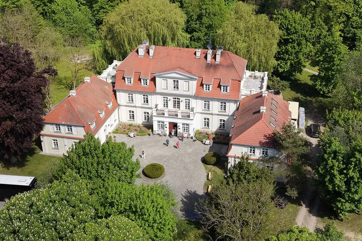 Malowniczy pałac Godętowo otoczony zielenią. Kilka osób spaceruje po dziedzińcu przed zabytkowym budynkiem.