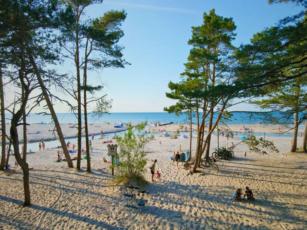 Słoneczna plaża w Dębkach z turystami, rowerami i strumieniem wpadającym do morza. Dębki promenada tętni życiem.