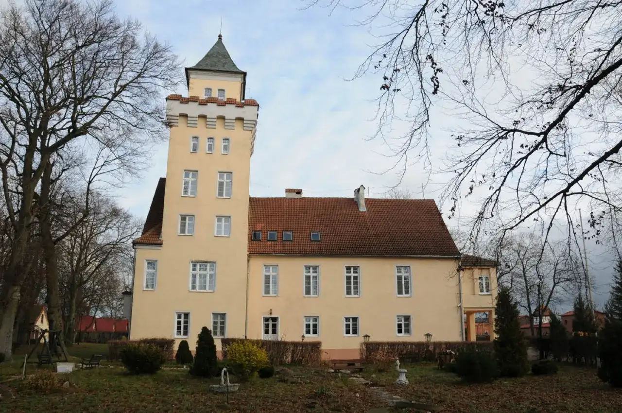Zamek Nowęcin, żółty budynek z wieżą i czerwonym dachem, otoczony drzewami i ogrodem.