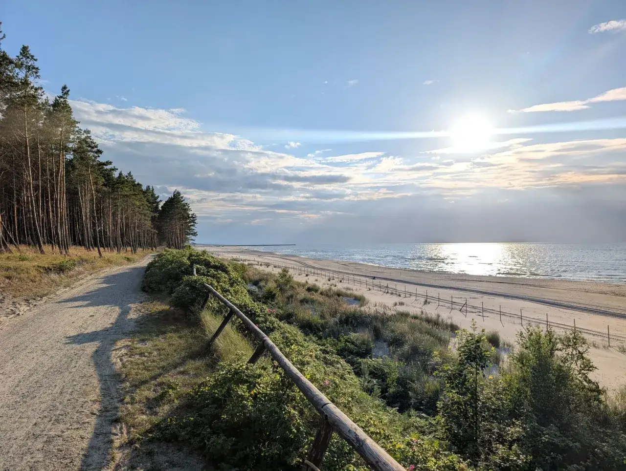 Piękna, piaszczysta plaża z lasem sosnowym po lewej. Słońce odbija się w morzu, tworząc błyszczącą ścieżkę. Idealna jantar trasa na spacer.