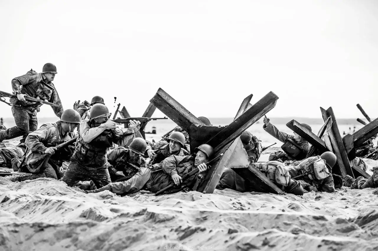 Żołnierze w hełmach i mundurach, z karabinami, ukrywają się za betonowymi fortyfikacjami na plaży. Scena przypomina D-Day, hel.