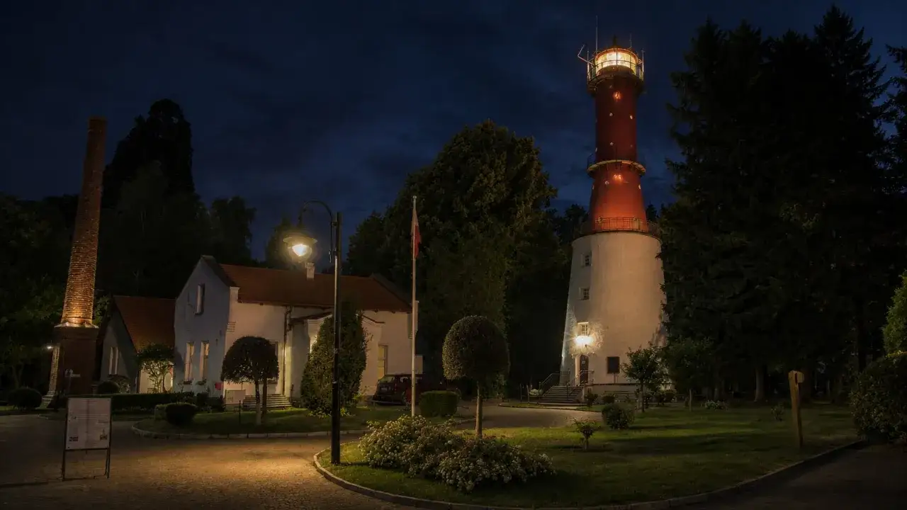 Nocna iluminacja latarni morskiej Rozewie. Obok niej stoją budynki i ceglany komin.