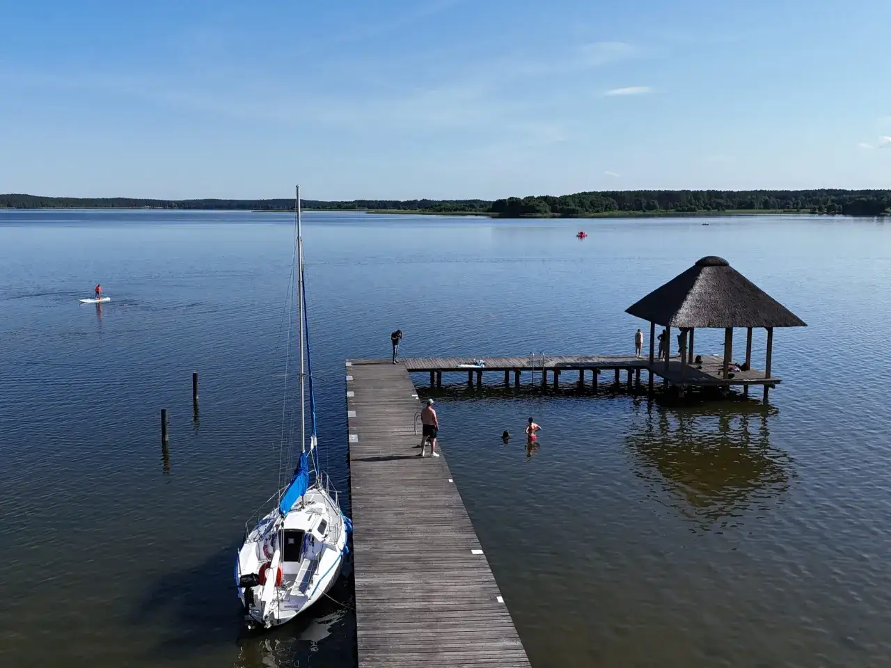 Letni dzień w Swornegaciach na Kaszubach. Drewniane molo, żaglówka, kąpiel w jeziorze i altana z trzciny.