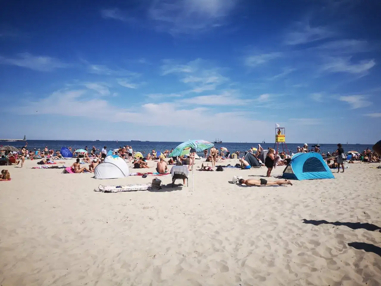 Tłum ludzi odpoczywa na piaszczystej plaży w Gdyni. Widać namioty, parasole i ratownika na wieży. Słoneczny dzień sprzyja plażowaniu na kąpieliskach Gdyni.