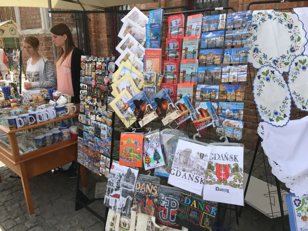 Stoisko z pamiątkami na targu w Gdańsku: magnesy, pocztówki, kubki i haftowane serwetki z motywami miasta.