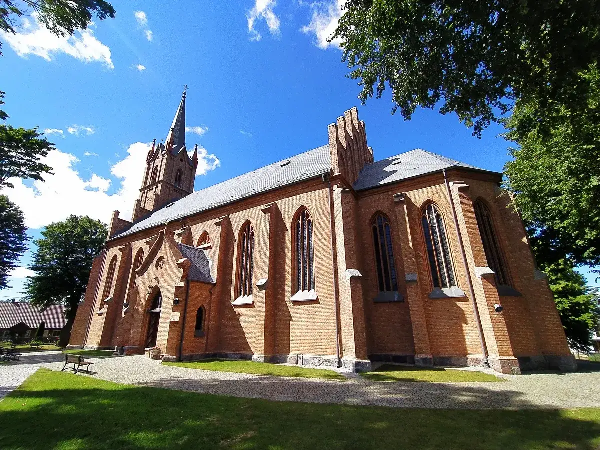 Swarzewo sanktuarium, ceglany kościół z wieżą i strzelistymi oknami, otoczony zielenią, pod błękitnym niebem z chmurami.