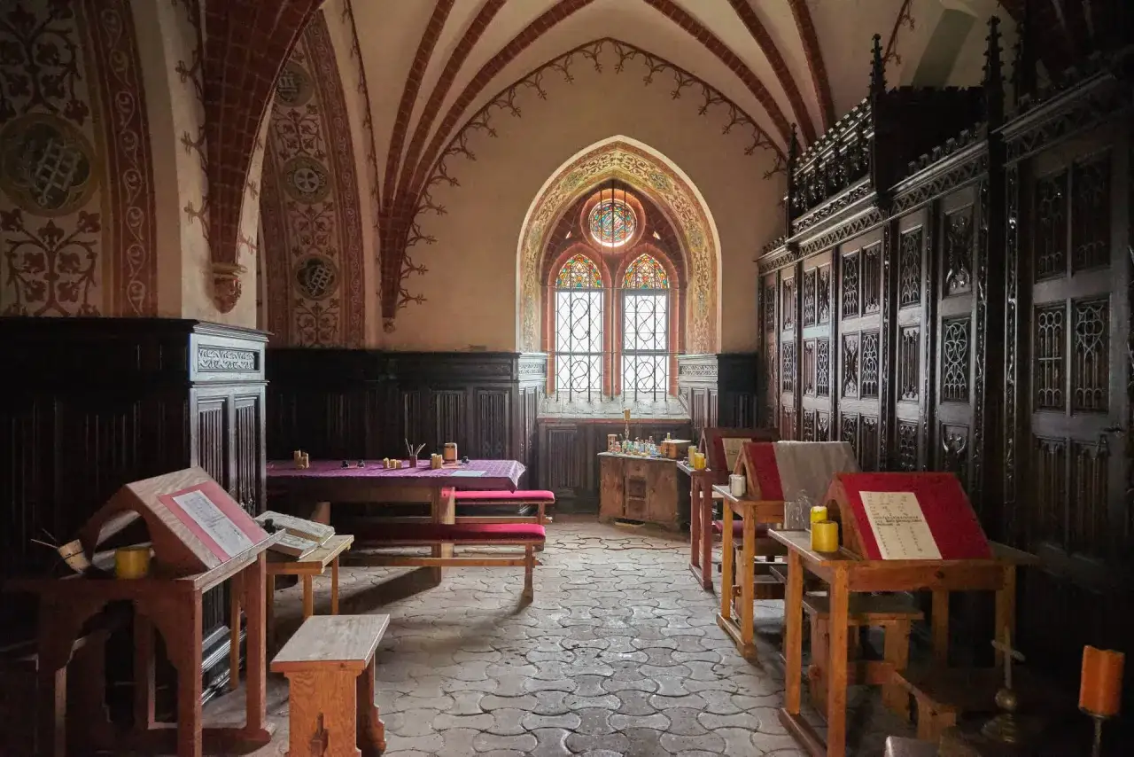 Wnętrze z gotyckimi łukami i witrażowym oknem. W sali znajdują się drewniane meble, stoły z księgami i ławki, tworząc atmosferę historycznych atrakcji Pelplina.