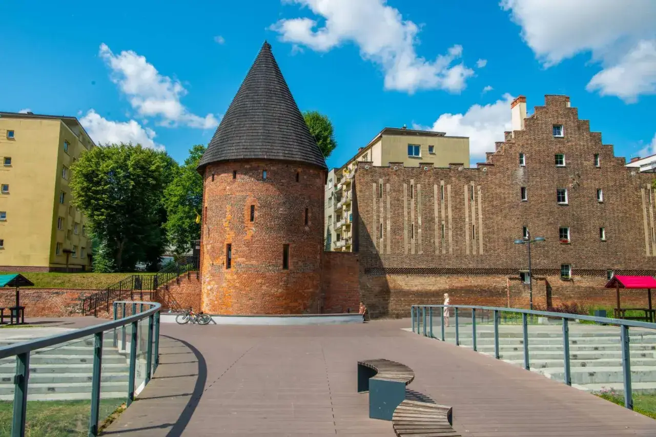 Kamienna baszta czarownic z stożkowym dachem, otoczona nowoczesną architekturą i zielenią, pod błękitnym niebem.