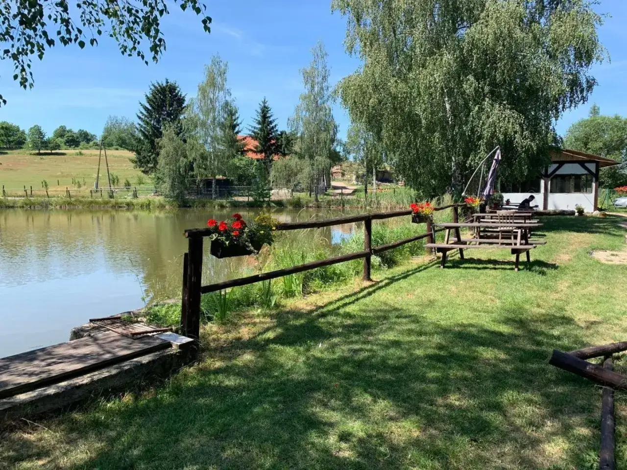 Drewniane ławki i stoły piknikowe nad spokojnym jeziorem, z kwitnącymi czerwonymi kwiatami na płocie. Idealne miejsce na odpoczynek nad potokiem grzędy.