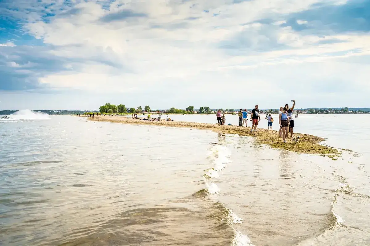 Ludzie spacerują po piaszczystym cyplu, tworzącym naturalną plażę w Rzucewie. W tle widać wodę i chmury.