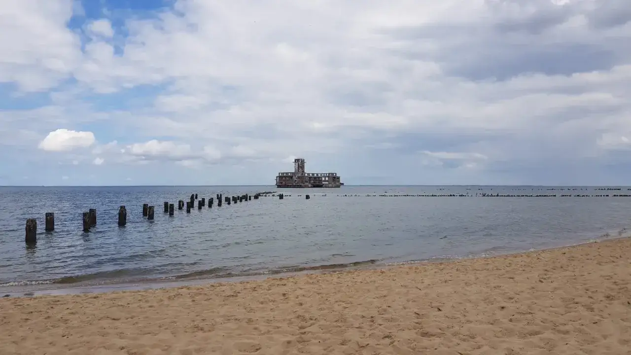 Betonowa torpedownia Hel, samotnie stojąca na morzu, otoczona drewnianymi słupkami.