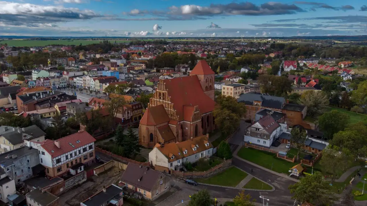 Widok z lotu ptaka na miasto z dominującym ceglanym kościołem w Pucku. Dachy dom&oacute;w i zieleń tworzą malowniczy krajobraz.