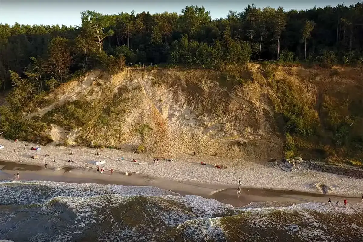 Jastrzębia G&oacute;ra, gdzie klif niczym gwiazda p&oacute;łnocy wznosi się nad plażą. Ludzie spacerują po piasku, fale obmywają brzeg.
