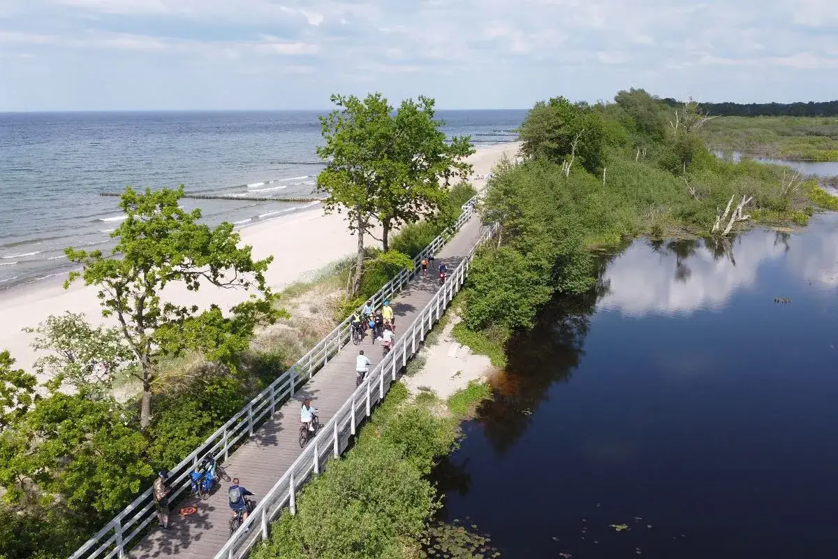 Rowerzyści przemierzają malowniczą trasę Velo Baltica R10, mijając plażę i spokojne wody jeziora.
